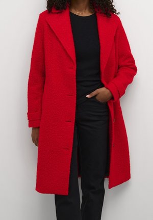 Vrouw die een felrode lange wollen jas draagt over een zwart shirt en zwarte broek, met één hand in haar zak.