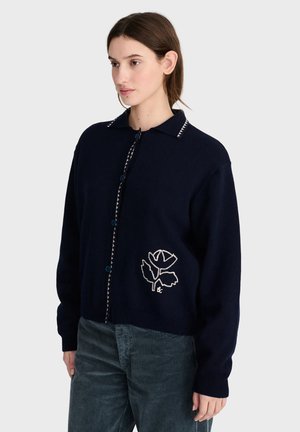 Femme portant un cardigan boutonné bleu marine foncé avec une fleur blanche brodée sur le côté et un pantalon texturé foncé, debout devant un fond uni.