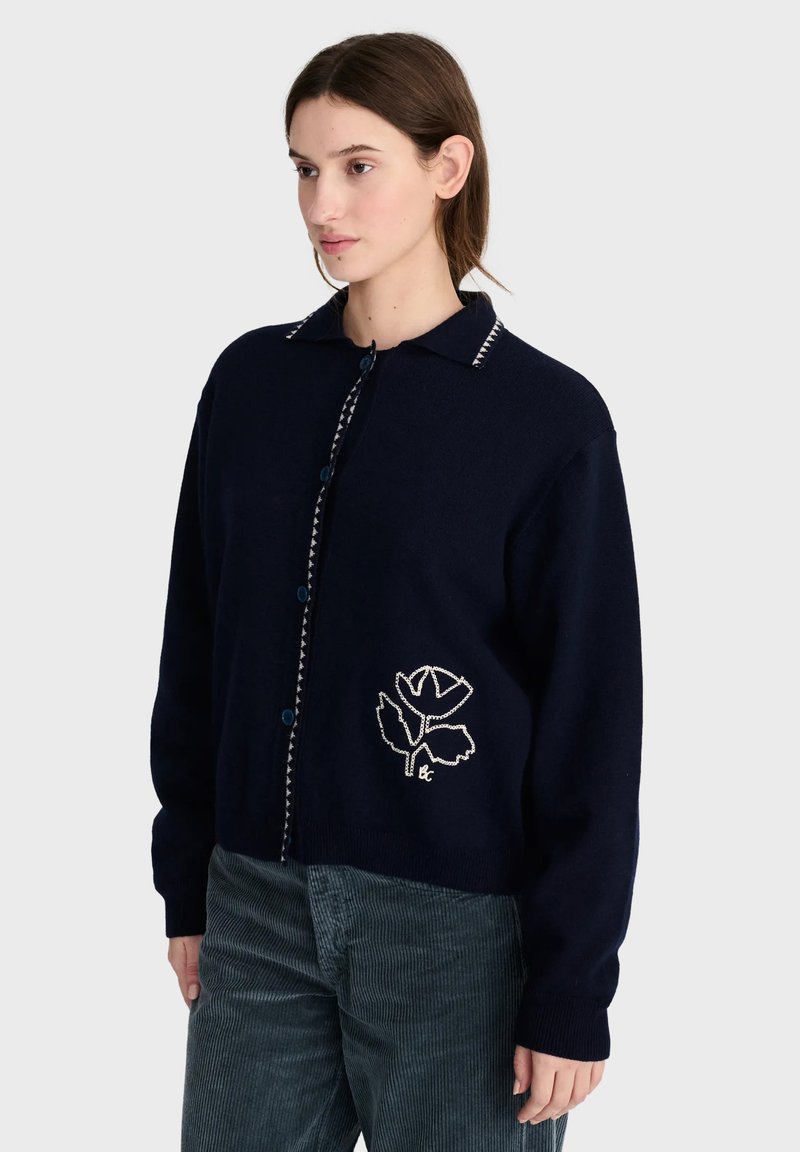 Femme portant un cardigan boutonné bleu marine foncé avec une fleur blanche brodée sur le côté et un pantalon texturé foncé, debout devant un fond uni.