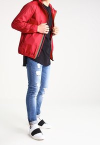 Chaqueta ligera roja con cremallera y capucha, llevada sobre una camiseta negra. Unos vaqueros azules desgastados y zapatillas blancas con tiras negras completan el conjunto.
