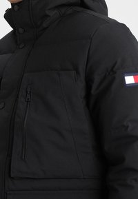 Černá prošívaná bunda s vysokým límcem, kapsou na zip na hrudi a logo nášivkou v barvách červené, bílé a modré na rukávu. Hladká textury.