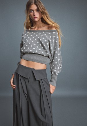 Femme aux longs cheveux portant un pull gris à pois décolleté et un pantalon large taille haute gris sur un fond gris uni.