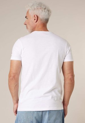 Homme d'âge moyen aux cheveux gris portant un t-shirt blanc uni et un jean en denim bleu clair, vu de dos sur un fond neutre.