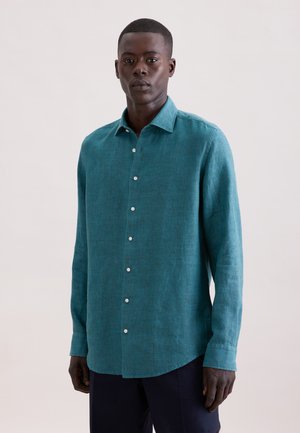 Homme debout portant une chemise à manches longues turquoise avec des boutons et un pantalon foncé, sur un fond clair uni.