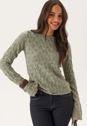 Kvinde smilende, iført en tekstureret lysegrøn langærmet top og mørke jeans, med halskæde og løst bølget hår, hånd på hoften.
