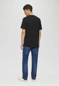 Schwarzes Baumwoll-T-Shirt, Rundhalsausschnitt, kurze Ärmel, lockere Passform, kombiniert mit blauen Denim-Jeans und weißen Sneakers, von hinten betrachtet.