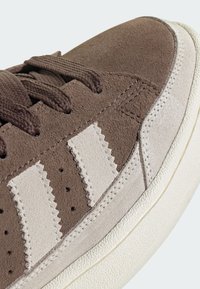 Brown suede sneaker with cream rubber sole. Features textured laces, three light beige stripes, and a smooth toe cap.