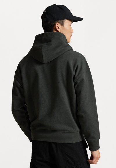 Σκούρο πράσινο φούτερ με κουκούλα και κορδόνι,  ribbed μανσέτες και στρίφωμα, υφή υφάσματος. Φορεμένο με μαύρο καπέλο, που δείχνει έναν μινιμαλιστικό σχεδιασμό.