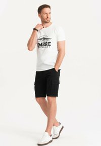 T-shirt bianca in cotone con grafica "AMBRE UNIVERSITY", abbinata a pantaloni cargo neri e sneakers bianche con dettagli marroni.