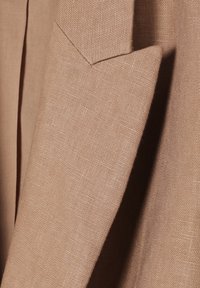 Tissu marron clair avec une apparence texturée et tissée. Présente un col pointu et des lignes épurées, avec des accents de couture visibles.