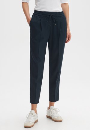 Personne portant un pantalon bleu marine avec cordon de serrage et plis à l'avant, et des baskets blanches, debout avec les mains partiellement dans les poches sur fond blanc.