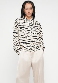 Chemise à manches longues beige avec rayures noires de tigre, dotée d'un col et d'une fermeture à boutons. Associée à un pantalon clair et ajusté.