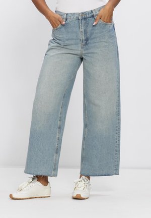 Persoon die lichtblauwe jeans met wijde pijpen en witte sneakers draagt, staand met handen in de zakken tegen een effen achtergrond.