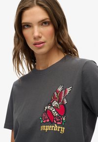 Camiseta gráfica de algodón en gris oscuro, con un corazón rojo con alas, rosas y la palabra "leal" en un diseño bordado.