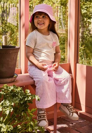 Young girl wearing a pink hat, beige t-shirt, and pink pants sits smiling on a wooden ledge in a sunlit garden setting.