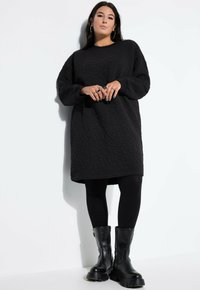 Vestido negro acolchado de gran tamaño con mangas largas, escote redondo y tejido texturizado, combinado con leggings negros y botas negras gruesas.