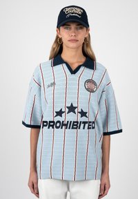 Jeune femme portant un pull tricoté bleu clair avec col bleu marine et le texte « PROHIBITED », motif rayé, et une casquette bleu marine avec texte blanc.