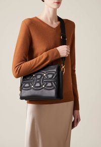 Black leather shoulder bag with quilted circular design, gold accents, and an adjustable strap. Model wears a brown sweater and satin skirt.