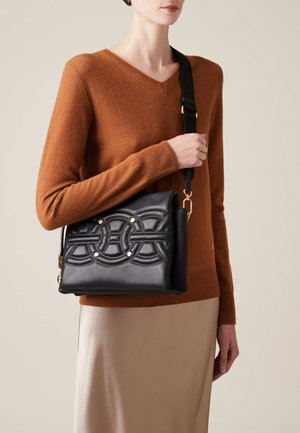 Black leather shoulder bag with quilted circular design, gold accents, and an adjustable strap. Model wears a brown sweater and satin skirt.