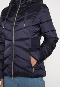 Giubbotto puffer blu navy con zip frontale, pannello laterale a coste, cuciture a chevron e accessori in tonalità oro. Tessuto liscio e lucido con tasche.