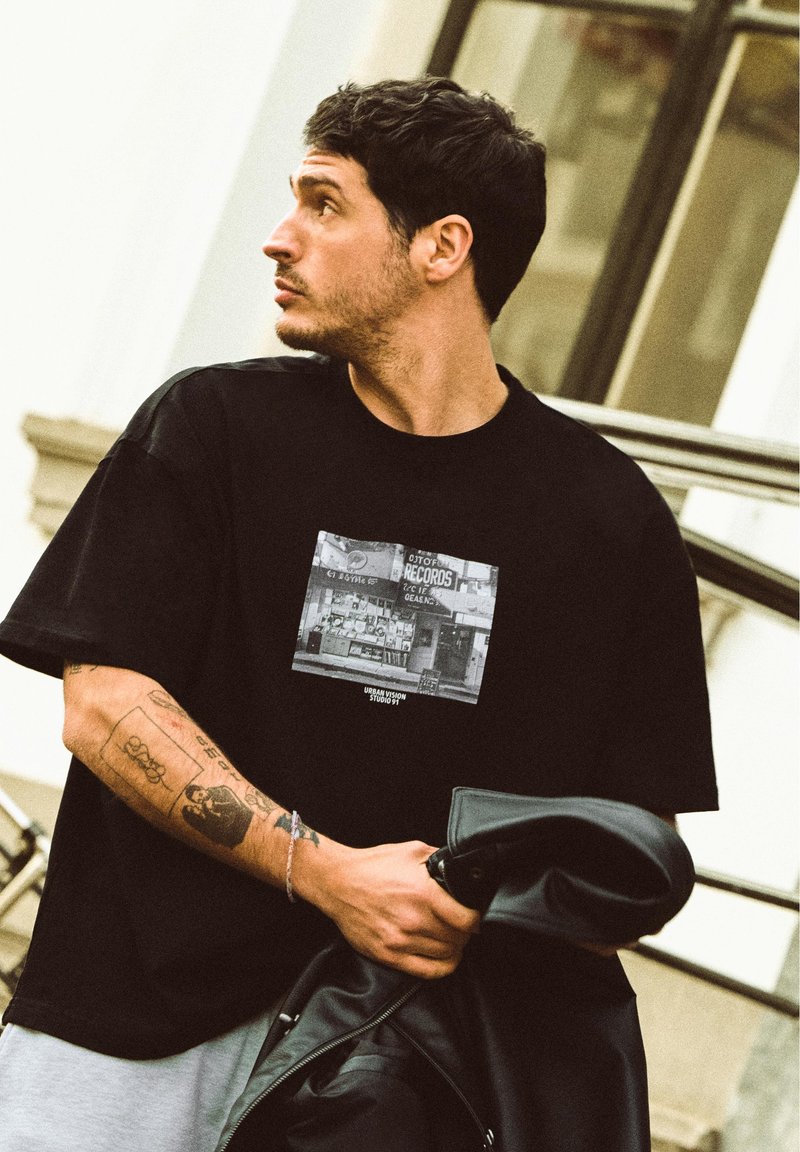 Homme avec des tatouages portant un t-shirt graphique noir tient une veste noire, regardant vers la gauche, debout à l'extérieur près d'un bâtiment moderne.