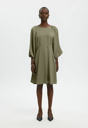 Femme debout portant une robe verte olive mi-longue avec de longues manches bouffantes et des talons noirs pointus sur fond blanc uni.