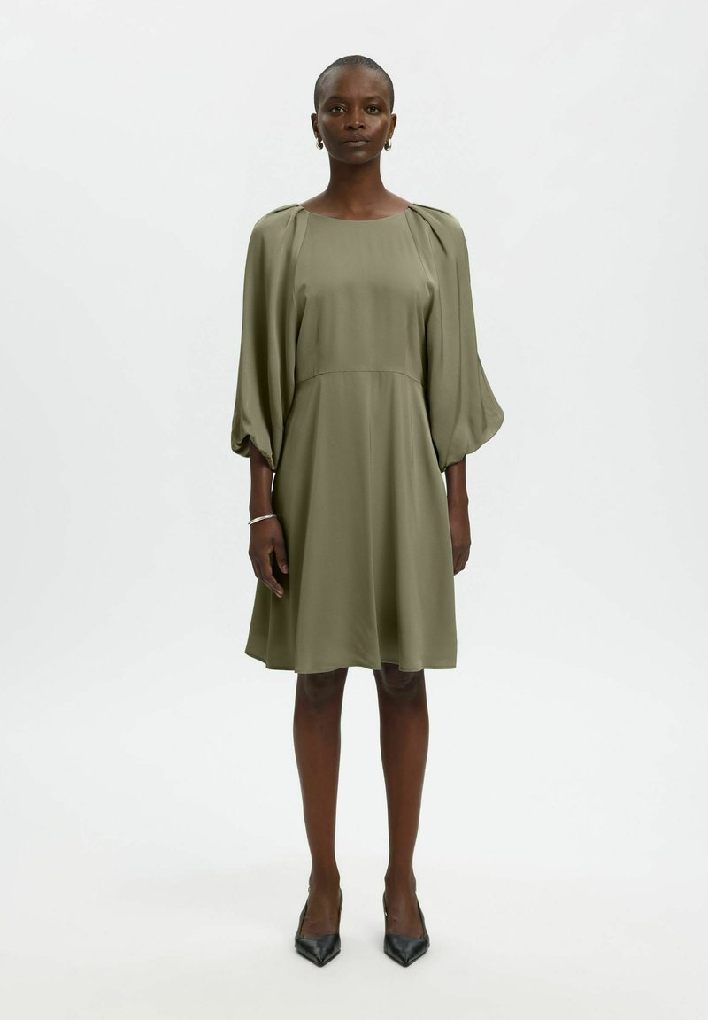Femme debout portant une robe verte olive mi-longue avec de longues manches bouffantes et des talons noirs pointus sur fond blanc uni.
