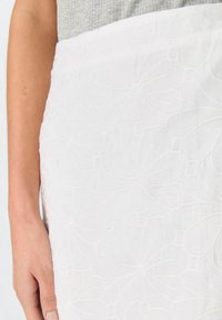 Falda blanca con un patrón floral bordado. Hecha de una tela texturizada. Presenta una cintura ajustada y un ligero brillo. Mano cerca del dobladillo.