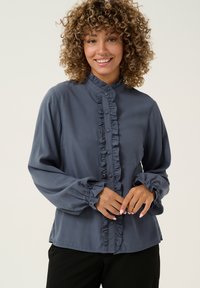 Blusa grigia con volant e colletto alto, con chiusura a bottoni e maniche lunghe a sbuffo. Tessuto morbido e liscio.