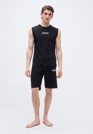 Young man standing barefoot wearing black sleeveless shirt and shorts with "GUESS" logo on chest and left thigh.