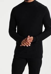Černý pletený svetr s kulatým výstřihem, manžetami a lemem s žebrováním. Nosený s rukama složenýma vpředu, minimalistický design, texturovaná tkanina.