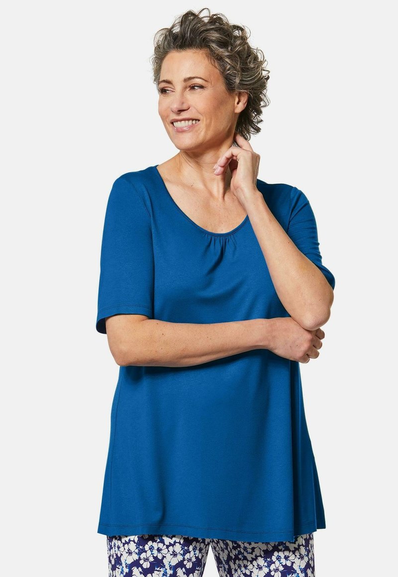 Blaues kurzärmliges Oberteil mit rundem Ausschnitt und gerafften Details, kombiniert mit floral gemusterten Hosen. Weicher, glatter Stoff.