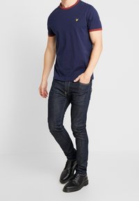 Mężczyzna w granatowej koszulce z czerwoną lamówką, ciemnych obcisłych dżinsach i czarnych skórzanych butach stojący na jednolitym białym tle.