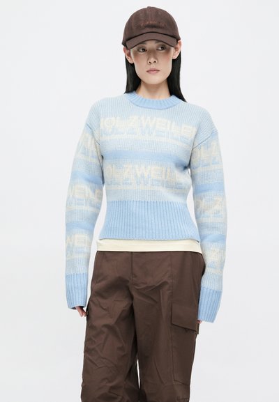 Šviesiai mėlynas nertos megztinis su kreminėmis horizontaliomis juostelėmis, kuriame yra tekstas. Nukirptas dizainas ir margas apdailos kraštas. Suderintas su rudos spalvos kargo kelnėmis.