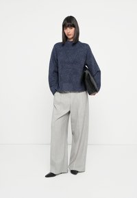 Marineblå strukturert genser med høy hals, kombinert med lysegrå vide bukser. Modellen holder en svart clutch, iført svarte spisse sko.