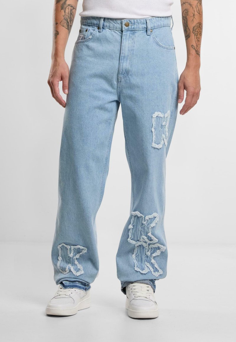 Lyseblå denimjeans med avslappet passform, med slitte lapper og broderte bokstaver på beina. Hvite joggesko fullfører looken.