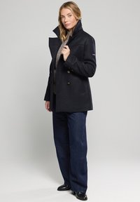 Abrigo de mezcla de lana en color navy con cuello puntiagudo, diseño de doble botonadura y botones de latón. Combinado con pantalones de denim de pierna ancha y calzado negro.