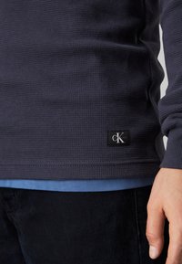 Maglia a maniche lunghe a coste navy con etichetta nera che presenta un logo bianco all'orlo, sovrapposta a una maglietta azzurro chiaro.