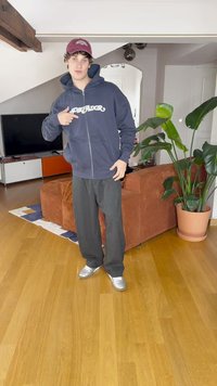 Marineblå zip-opp hettegenser med hvit "PREDADOR" tekst, grå vide bukser og sølvfargede joggesko, stående i en stue med en plante.