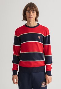 Rot und marine gestreifter Pullover mit geripptem Halsausschnitt und Bündchen, mit einem kleinen Logopatch auf der linken Brust. Weicher Strickstoff.