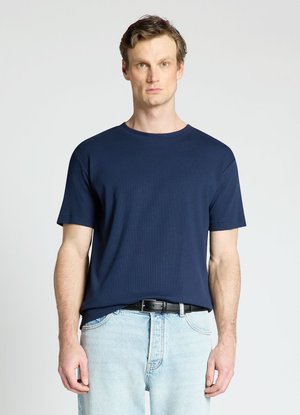 Uomo con capelli castani corti che indossa una maglietta a maniche corte blu navy infilata in jeans azzurri con una cintura nera.