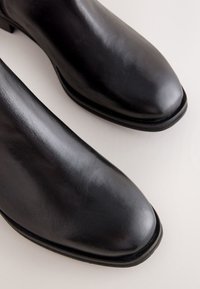 Paire de chaussures habillées en cuir noir poli à bouts arrondis exposée sur une surface claire.