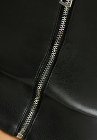 Svart faux läderplagg med en central dragkedja med silverfärgad tandning, slät textur och eleganta linjer som framhäver en åtsittande silhuett.