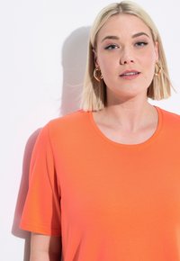Světle oranžové tričko s krátkým rukávem a kulatým výstřihem, hladká textura a jemný lesk. Zlaté kruhové náušnice jako výrazné doplňky.