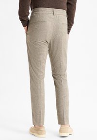 Man in beige geruite slim-fit broek, bruine trui en beige suede instappers, staande met handen in de zakken.