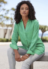 Green button-up shirt made of lightweight fabric, featuring long sleeves and a relaxed fit, paired with light gray jeans and a gold chain bracelet.