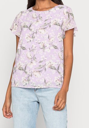Femme portant un chemisier lilas à manches courtes avec un motif floral blanc, associé à un jean en denim bleu clair, debout devant un fond uni.