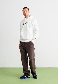 Hanorac alb cu glugă, având logo-ul Nike în negru și auriu, asociat cu pantaloni maro și încălțăminte neagră cu talpă albastră, stând pe un podea verde.