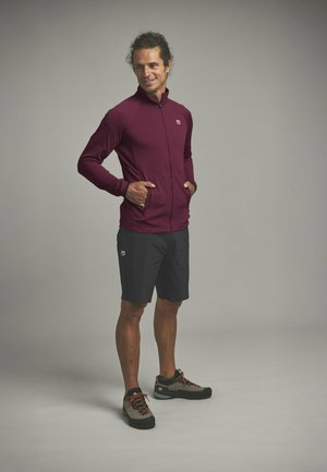 Mann steht mit den Händen in den Taschen, trägt eine burgunderrote Reißverschlussjacke, schwarze Shorts und graue Wanderschuhe vor einem einfarbigen Hintergrund.