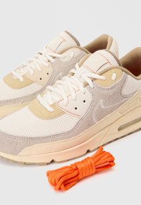 Béžové a biele tenisky Nike s bielymi šnúrkami a ďalším párom skrútených jasne oranžových šnúrok na bielom pozadí.